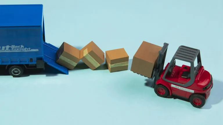 Un chariot élévateur de jouets déplace une boîte vers un camion de livraison de jouets bleu, avec trois boîtes dispersées entre eux sur un fond bleu clair, ressemblant à une scène de réalité augmentée.